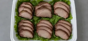 Open turn-in box with six portions of sliced smoked pork on a lettuce bed