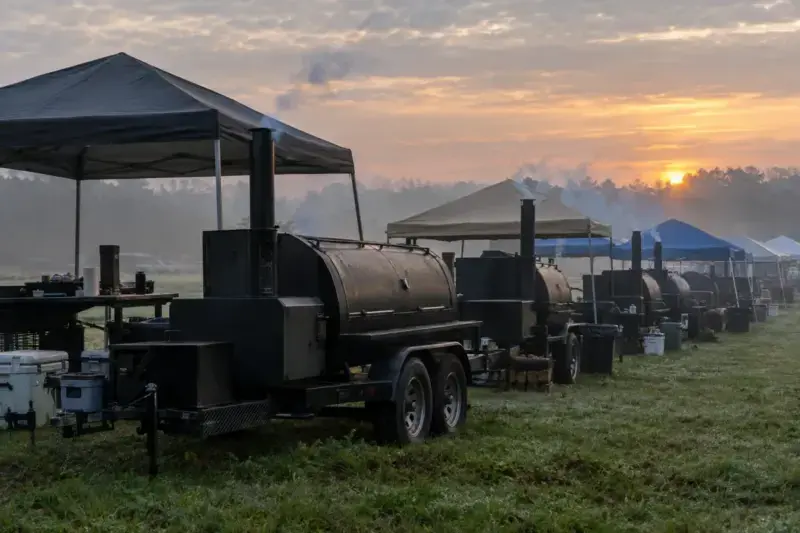Competition barbecue field at first light, offset smokers in a row with smoke rising